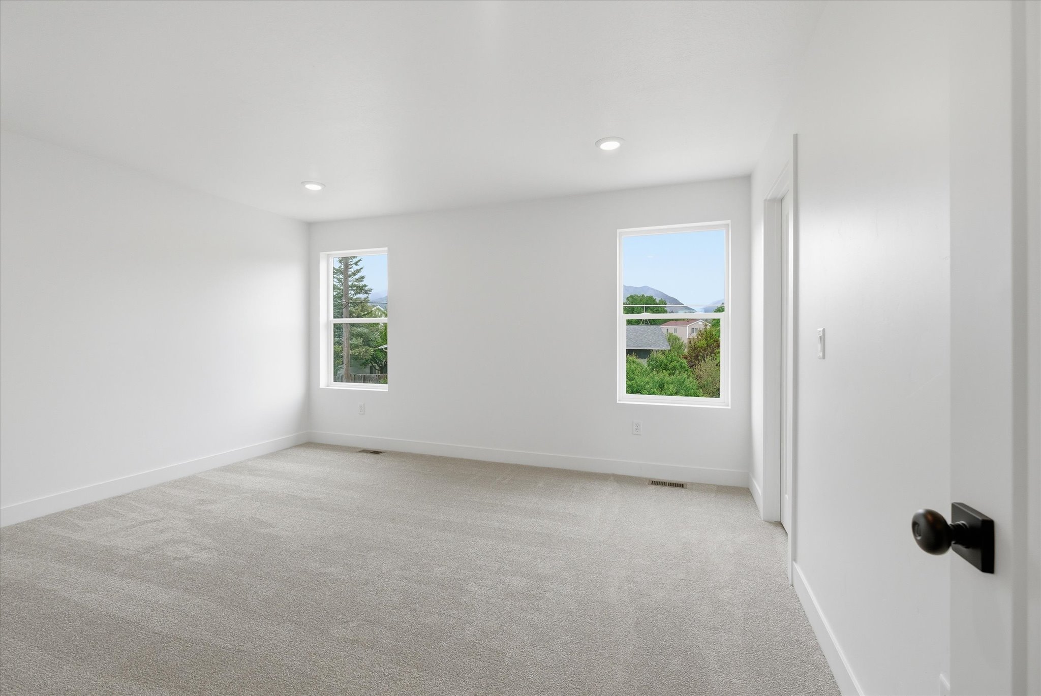 Spacious room featuring light gray carpeting, crisp white walls, recessed ceiling lighting, and two windows offering a mountain backdrop