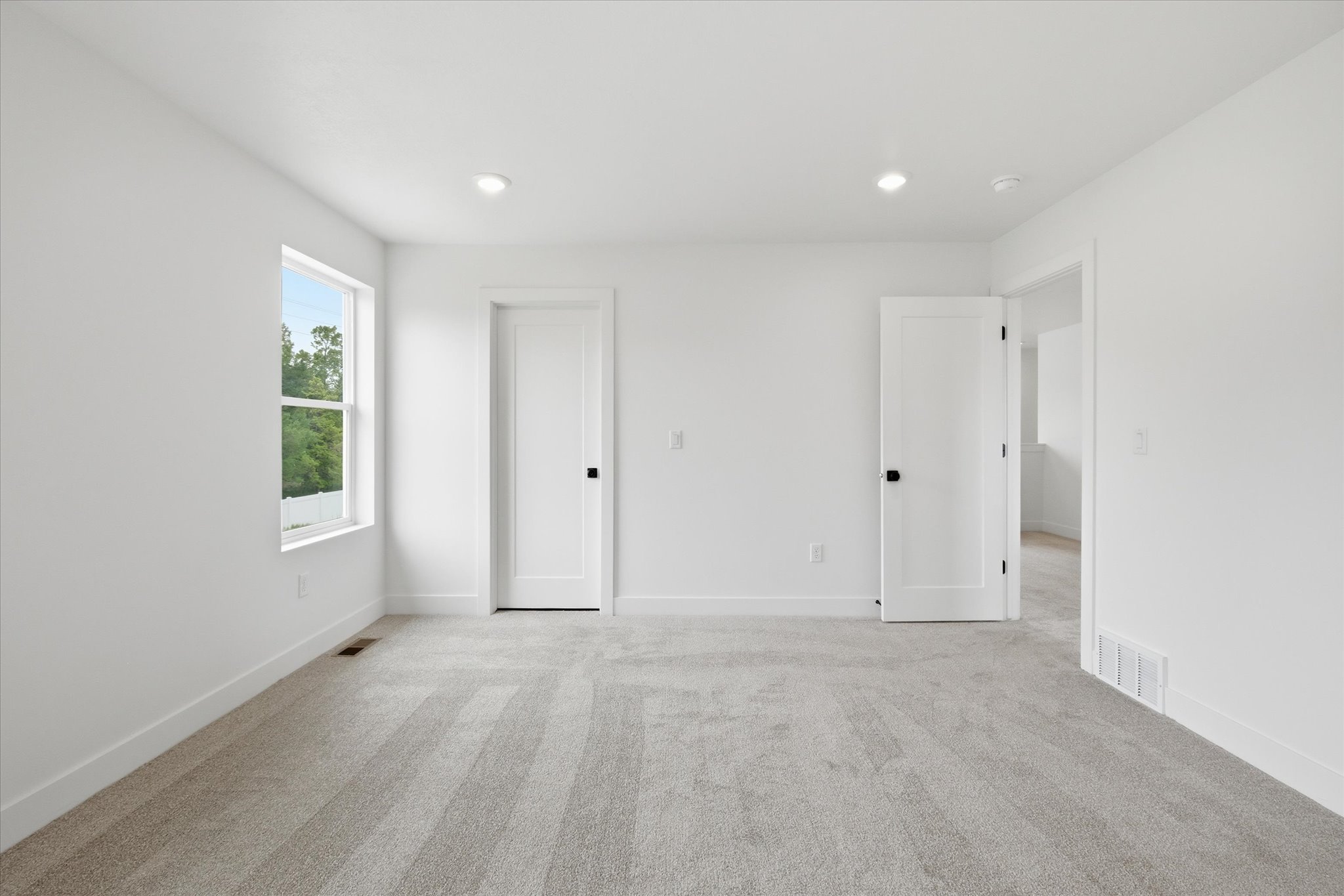 Spacious room featuring light gray carpet, white walls, two recessed ceiling lights, a white window with a green outdoor backdrop, and two white interior doors with black hardware