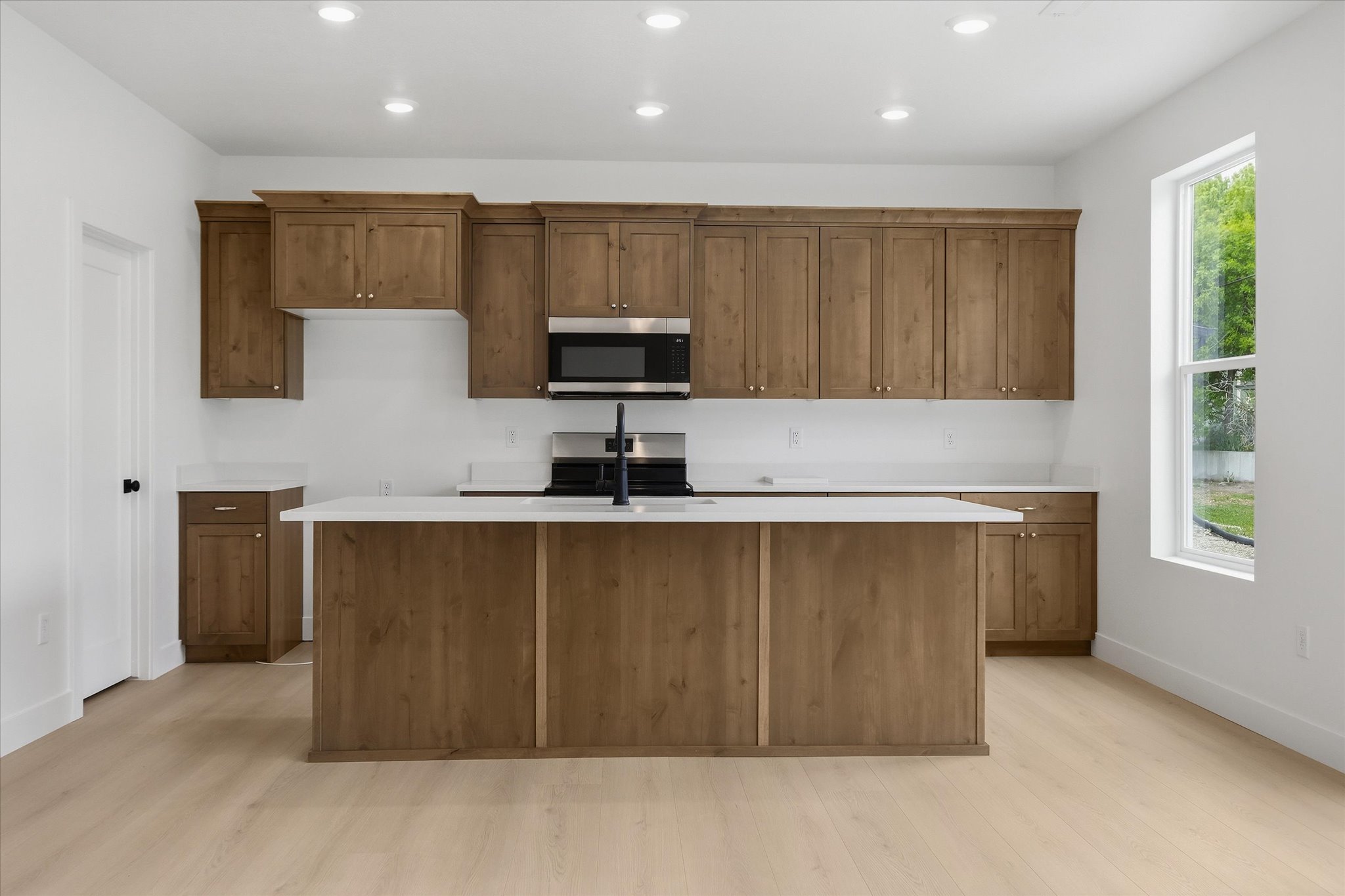 Kitchen featuring a central island with white countertop, extensive wood-finish cabinetry, stainless steel microwave, black cooktop, and light wood-finish flooring