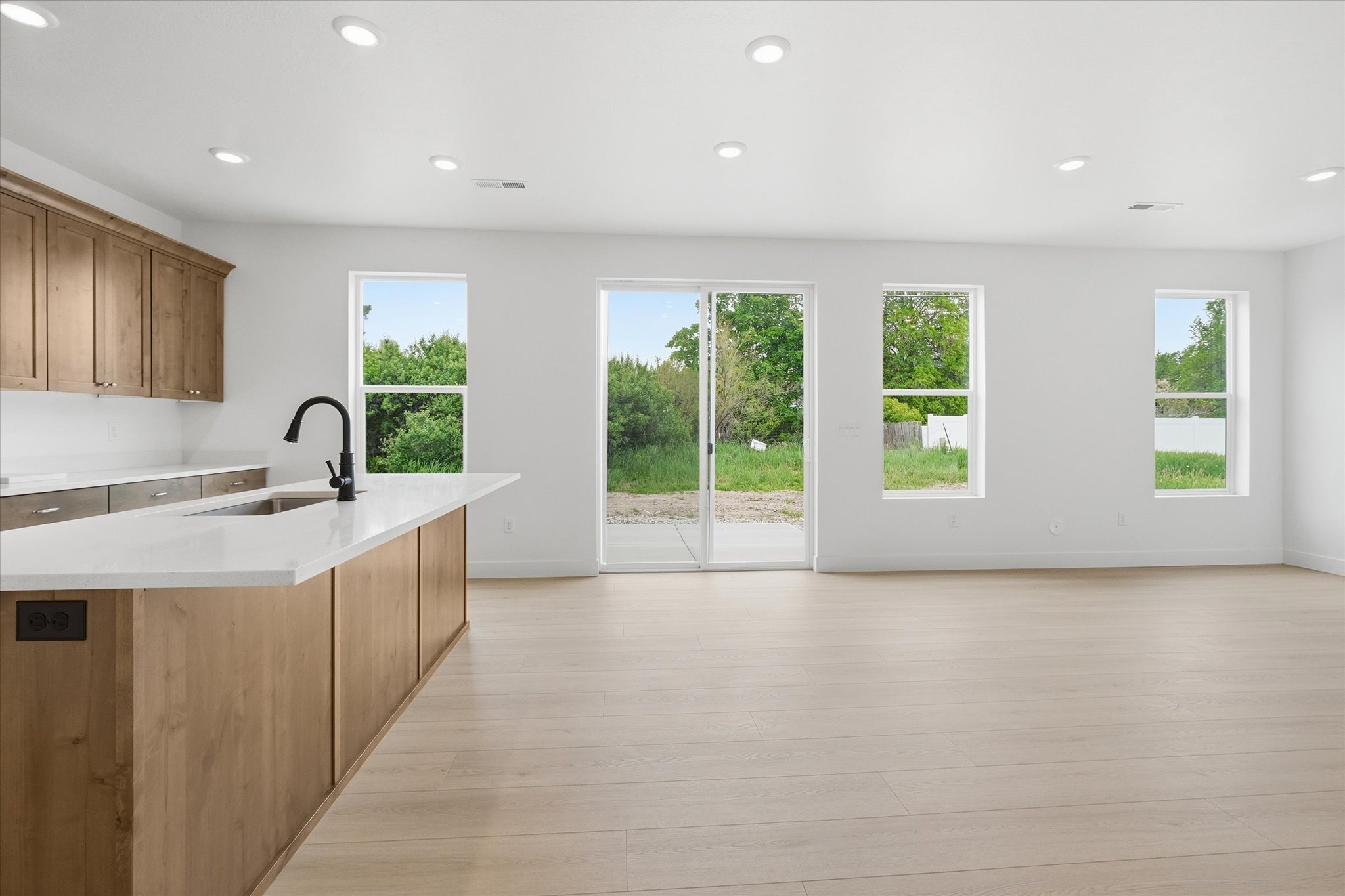 Open-concept living space with light wood-finish flooring, a kitchen island featuring a sink and matte black faucet, recessed lighting, and multiple windows providing natural illumination