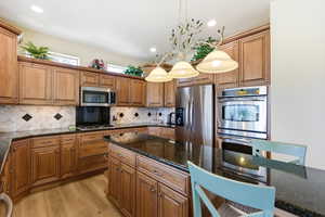 Kitchen with stainless steel appliances, wood finish cabinetry, dark stone counters, and decorative light fixtures