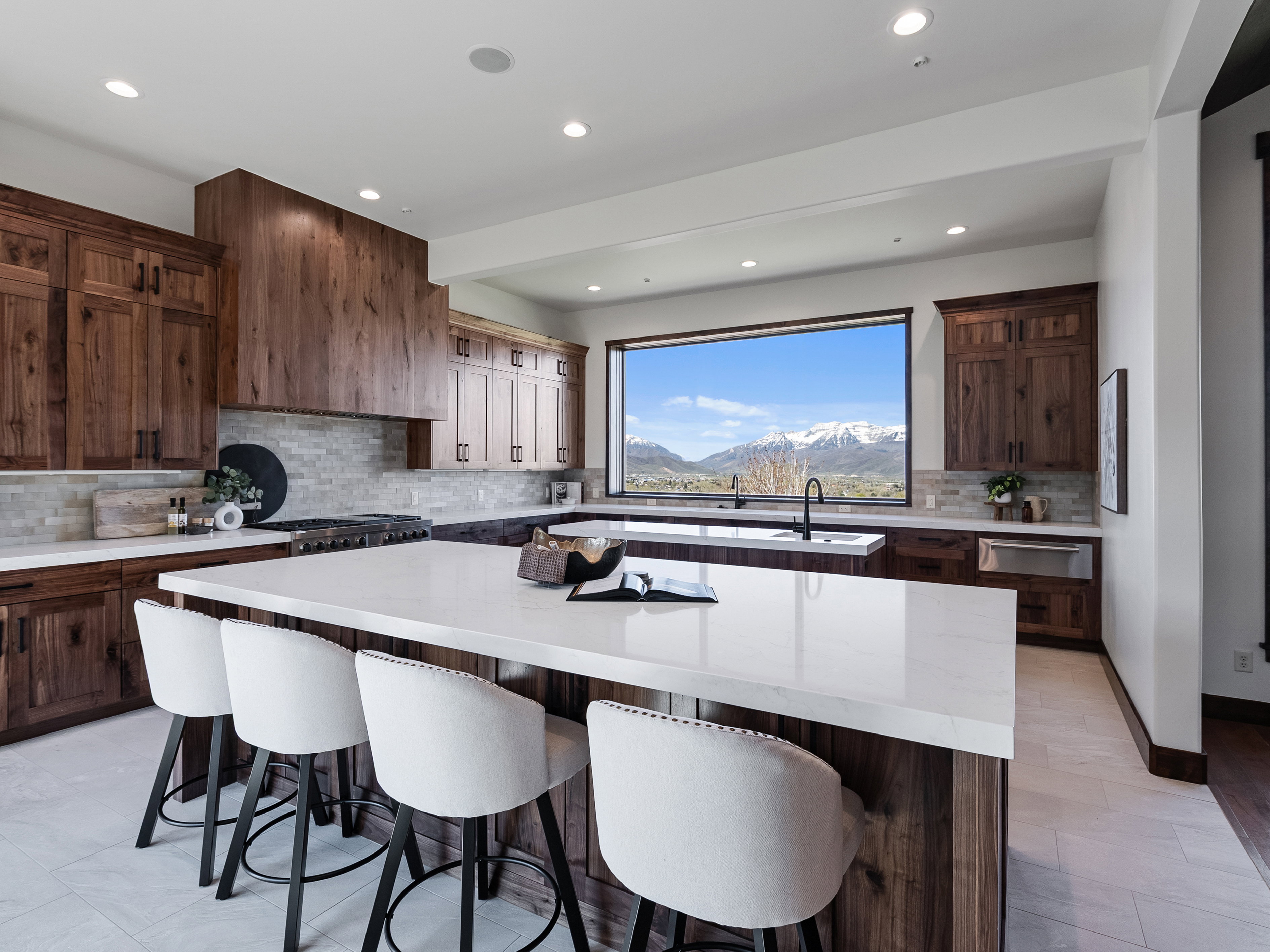 Kitchen featuring a substantial island with a light-toned countertop, a range with a wood-paneled hood, extensive wood cabinetry, and a large picture window showcasing a mountain landscape