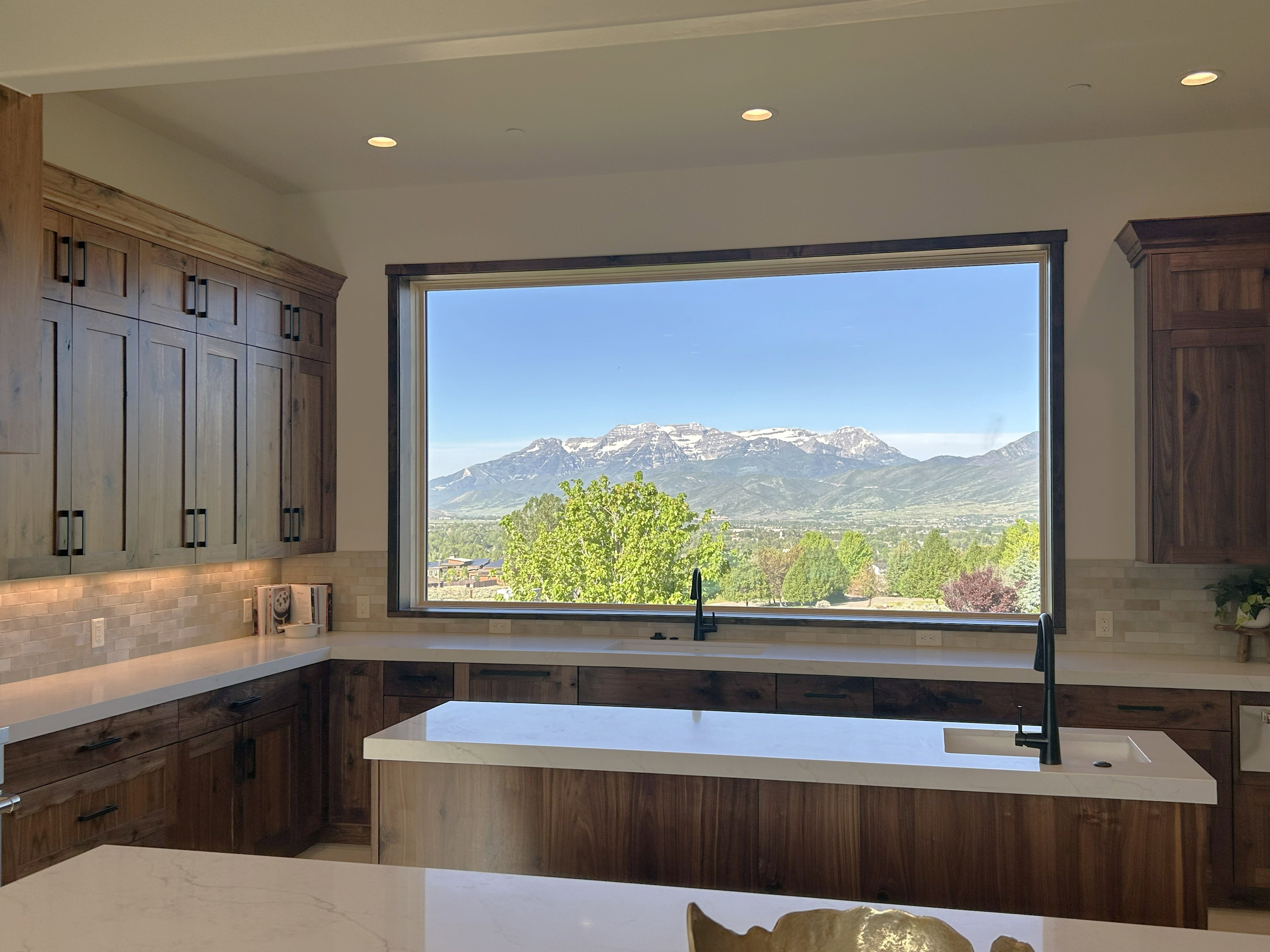 Kitchen featuring a large picture window with mountain and valley scenery, wood-finish cabinetry, light-toned countertops, tile backsplash, recessed lighting, and a kitchen island with a built-in sink