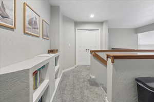Hallway with a textured wall, light colored carpet, and an upstairs landing