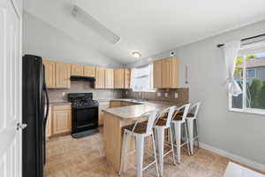 Kitchen with a breakfast bar, light wood finish cabinets, a peninsula, vaulted ceiling, and black appliances