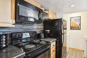 Kitchen with black appliances, light wood finish cabinetry, dark countertops, tasteful backsplash, and a textured ceiling