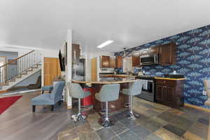 Kitchen with a kitchen breakfast bar, stainless steel appliances, wallpapered walls, dark wood finish cabinets, and dark stone finish flooring