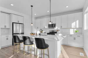 Kitchen featuring stainless steel appliances, white cabinets, light wood-style floors, an island with sink, and hanging light fixtures