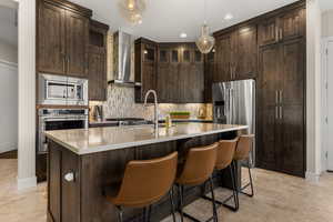 Kitchen featuring dark wood finish cabinets, stainless steel appliances, an island with sink, decorative backsplash, and decorative light fixtures