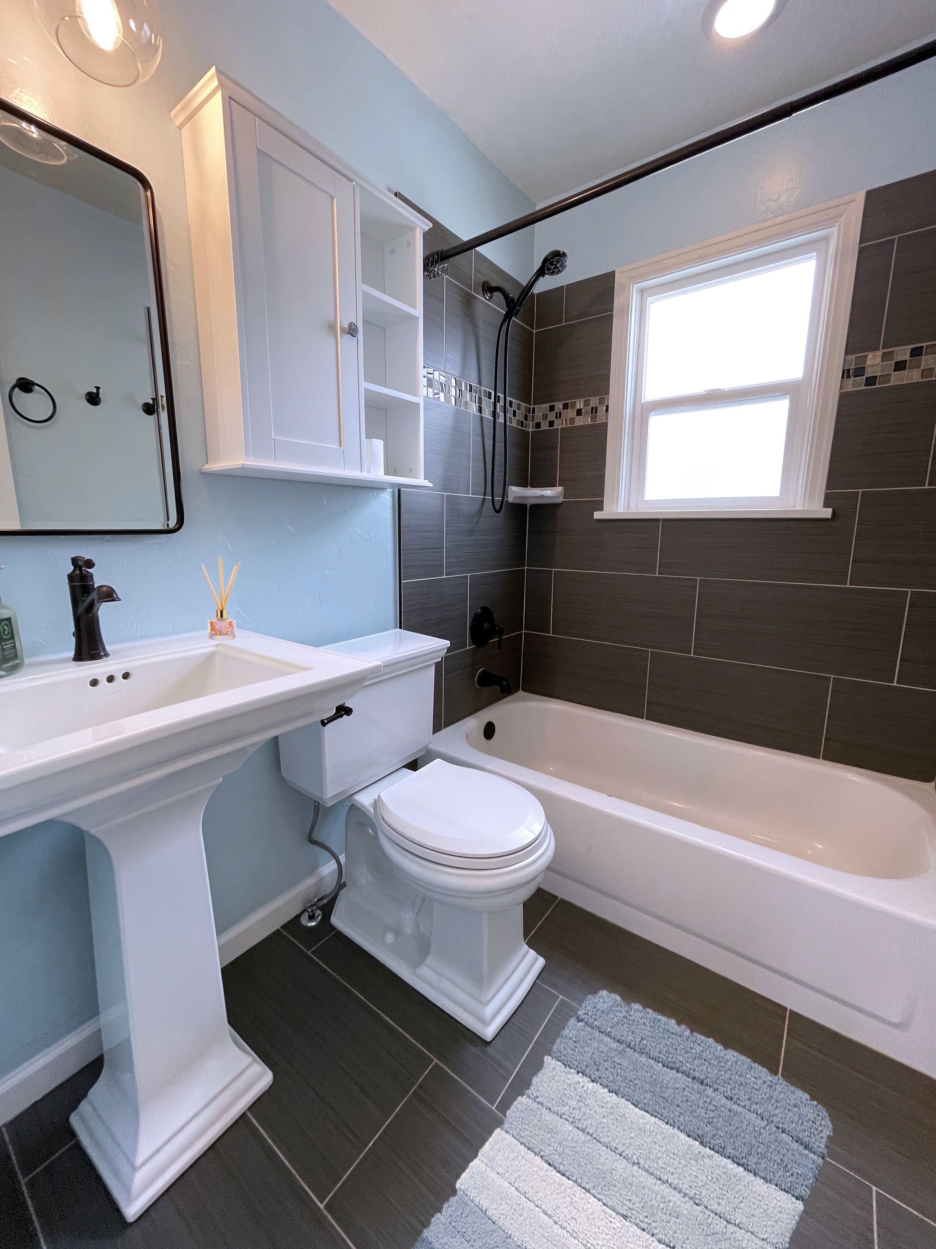 Contemporary bathroom featuring a pedestal sink, dark tile flooring, a tub/shower combination with dark tile surround, a built-in storage cabinet, and a window for natural light