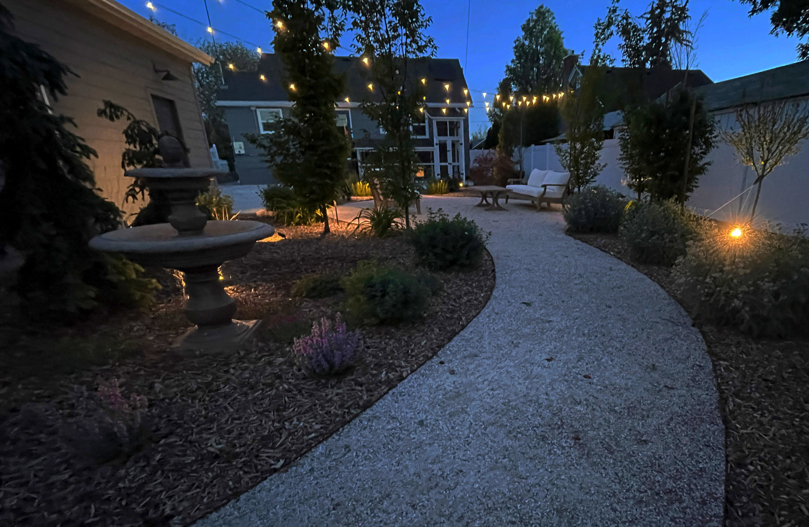 Gravel pathway leading to a landscaped garden featuring a tiered stone fountain, mature plantings, and string lighting