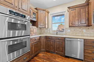 Kitchen featuring stainless steel appliances, dark wood-style floors, glass fronted cabinets, light stone countertops, and decorative backsplash