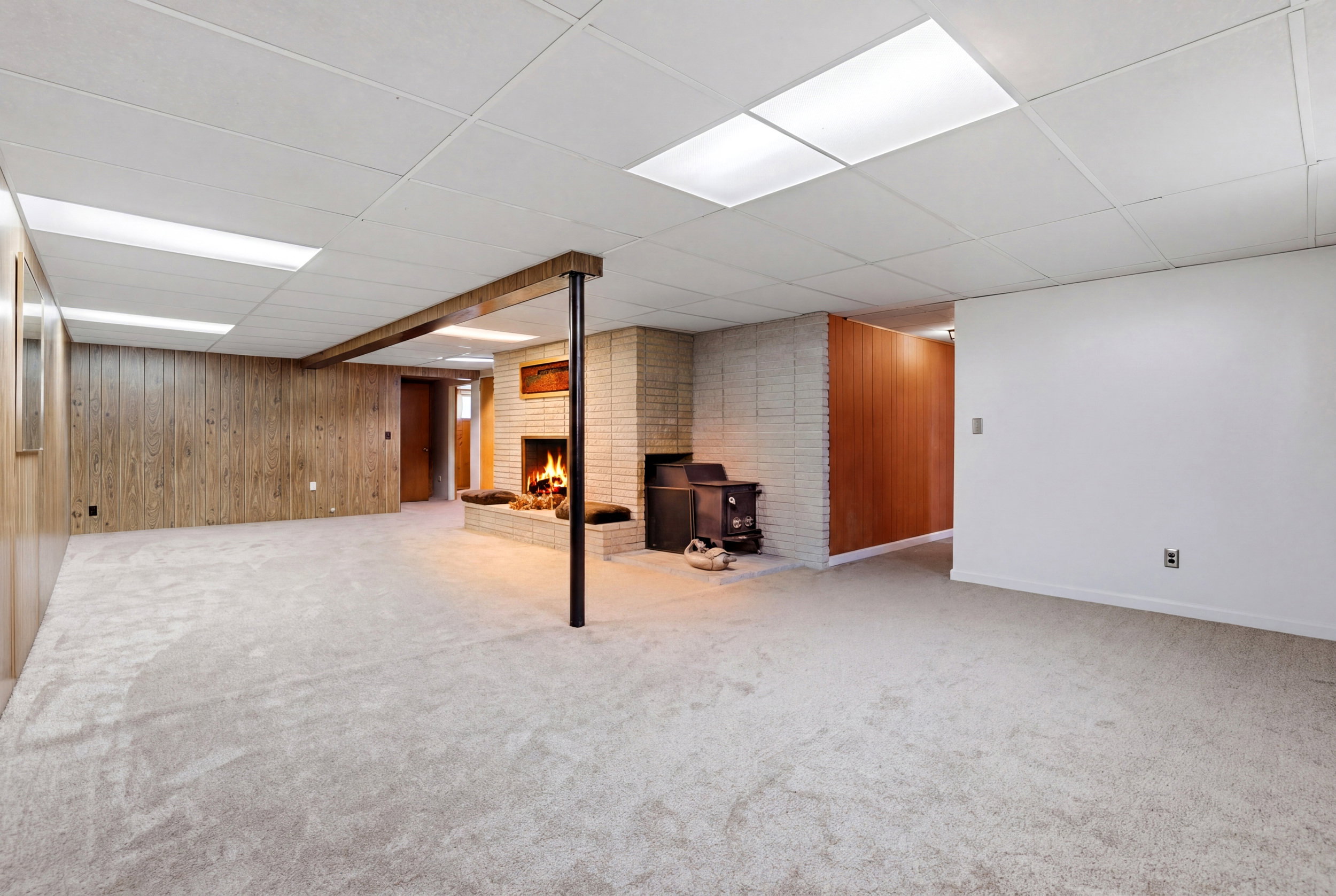 Finished basement featuring a brick fireplace, wooden walls, a drop ceiling, and light colored carpet