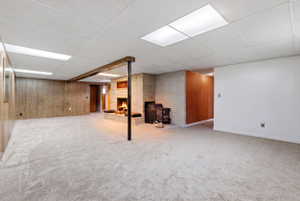 Finished basement featuring a brick fireplace, wooden walls, a drop ceiling, and light colored carpet