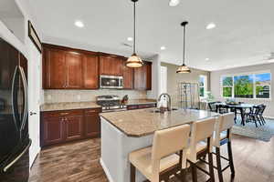 Kitchen with light stone countertops, a breakfast bar, hanging light fixtures over the island area, stainless steel appliances, and dark wood-type flooring