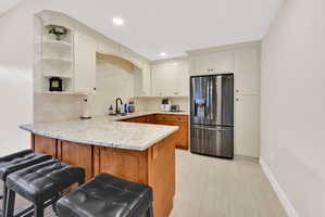 Dual tone kitchen featuring stainless steel refrigerator with ice dispenser, open shelves, a peninsula, a breakfast bar, and light stone countertops