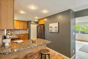 Kitchen featuring dark stone counters, stainless steel refrigerator with ice dispenser, a kitchen breakfast bar, wood finish cabinets, and recessed lighting