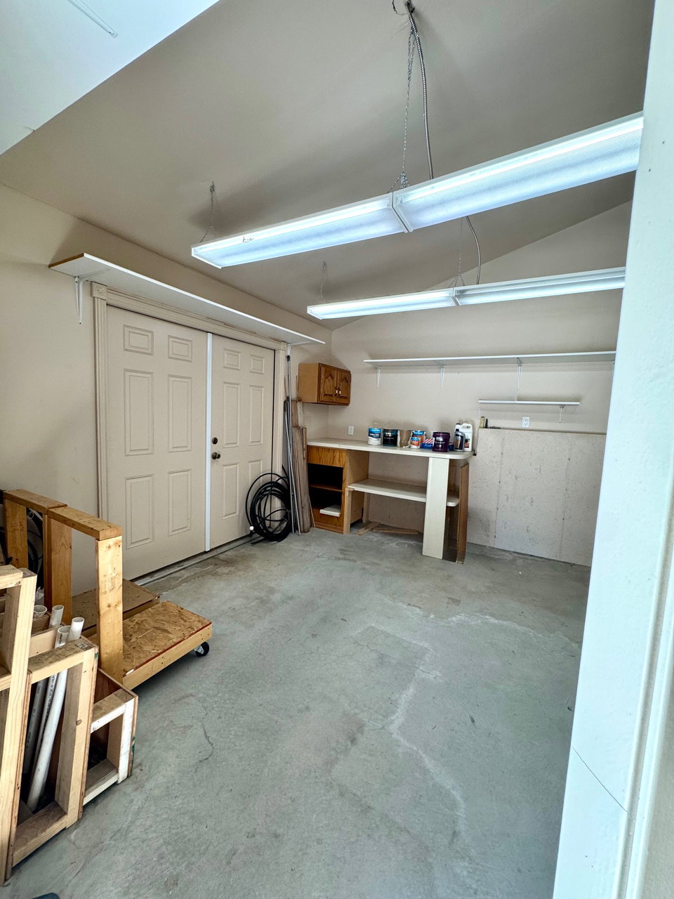 Spacious garage interior featuring a concrete floor, two fluorescent light fixtures, two built-in wall shelves, and a built-in workbench with a storage cabinet