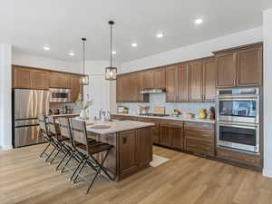 Kitchen with a breakfast bar, stainless steel appliances, pendant lighting, a center island with sink, and light wood-type flooring