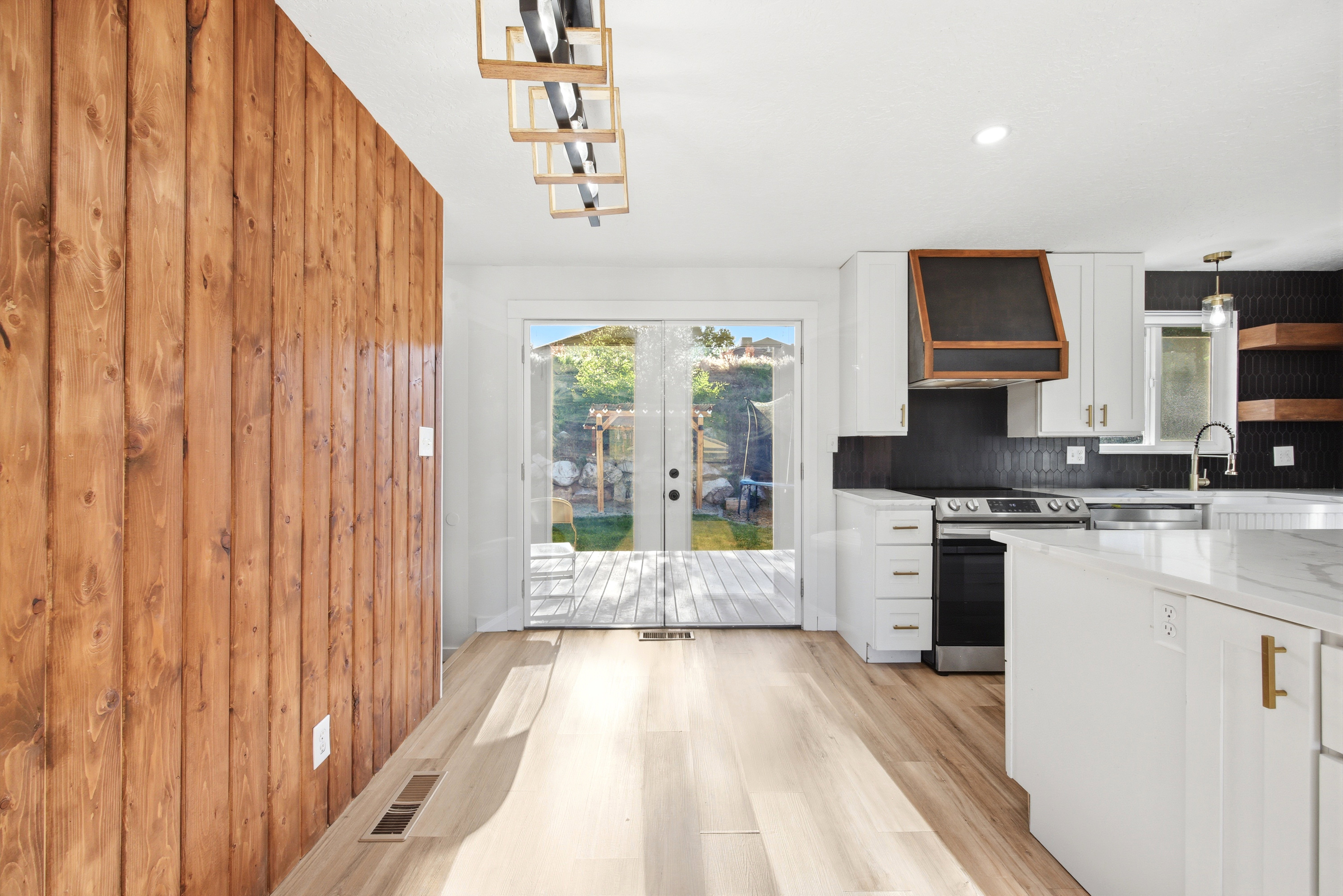 Open-concept kitchen and dining area featuring light wood-finish flooring, a wood-paneled accent wall, white cabinetry with gold hardware, a built-in range hood with wood trim, and double glass doors leading to an exterior deck