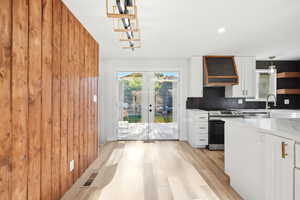 Open-concept kitchen and dining area featuring light wood-finish flooring, a wood-paneled accent wall, white cabinetry with gold hardware, a built-in range hood with wood trim, and double glass doors leading to an exterior deck
