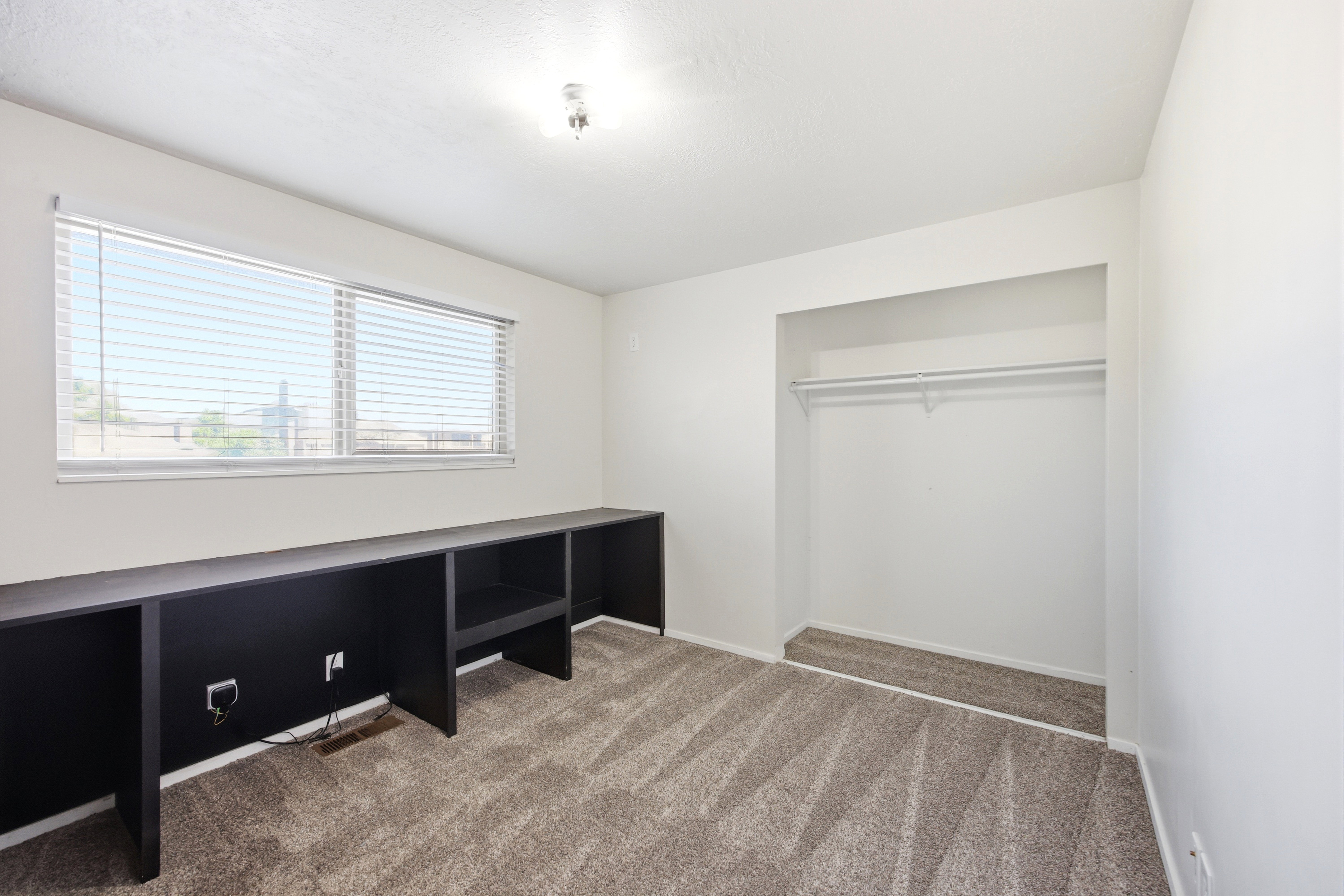 Room featuring a large window with blinds, a built-in dark storage and desk unit, wall-to-wall carpeting, and an open closet with a hanging rod
