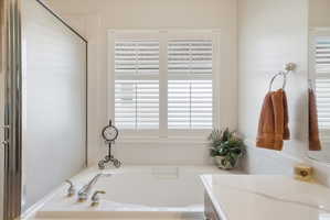 Soaking tub featuring polished chrome faucet with dual handles, white plantation shutters, frosted glass shower enclosure, and a stone-finish countertop