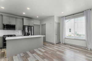 Kitchen featuring gray cabinets, stainless steel appliances, light wood-style flooring, an island with sink, and decorative backsplash