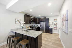 Kitchen featuring a kitchen bar, dark wood finish cabinetry, stainless steel appliances, light stone counters, and light wood-style floors