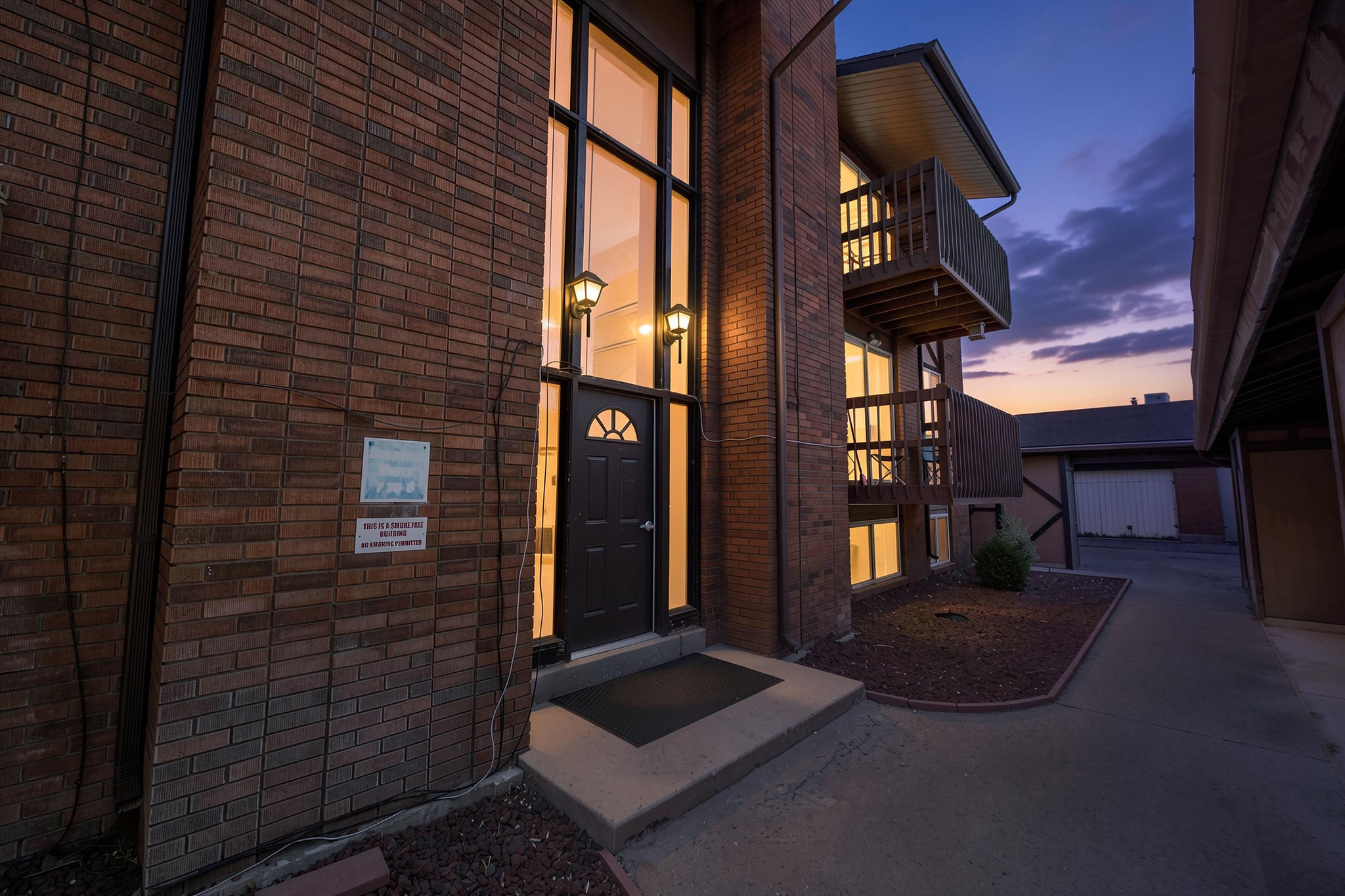 Brick facade building featuring a full-light entry door with an arched transom, exterior wall sconces, multi-level balconies with wood railings, and a concrete walkway