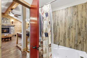 Bathroom with wood walls, wood-type flooring, a stone fireplace, a combined bath / shower with jetted tub, and lofted ceiling with beams