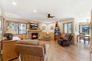 Living area featuring a wainscoted wall, a stone fireplace, a ceiling fan, wood walls, and light wood finished floors
