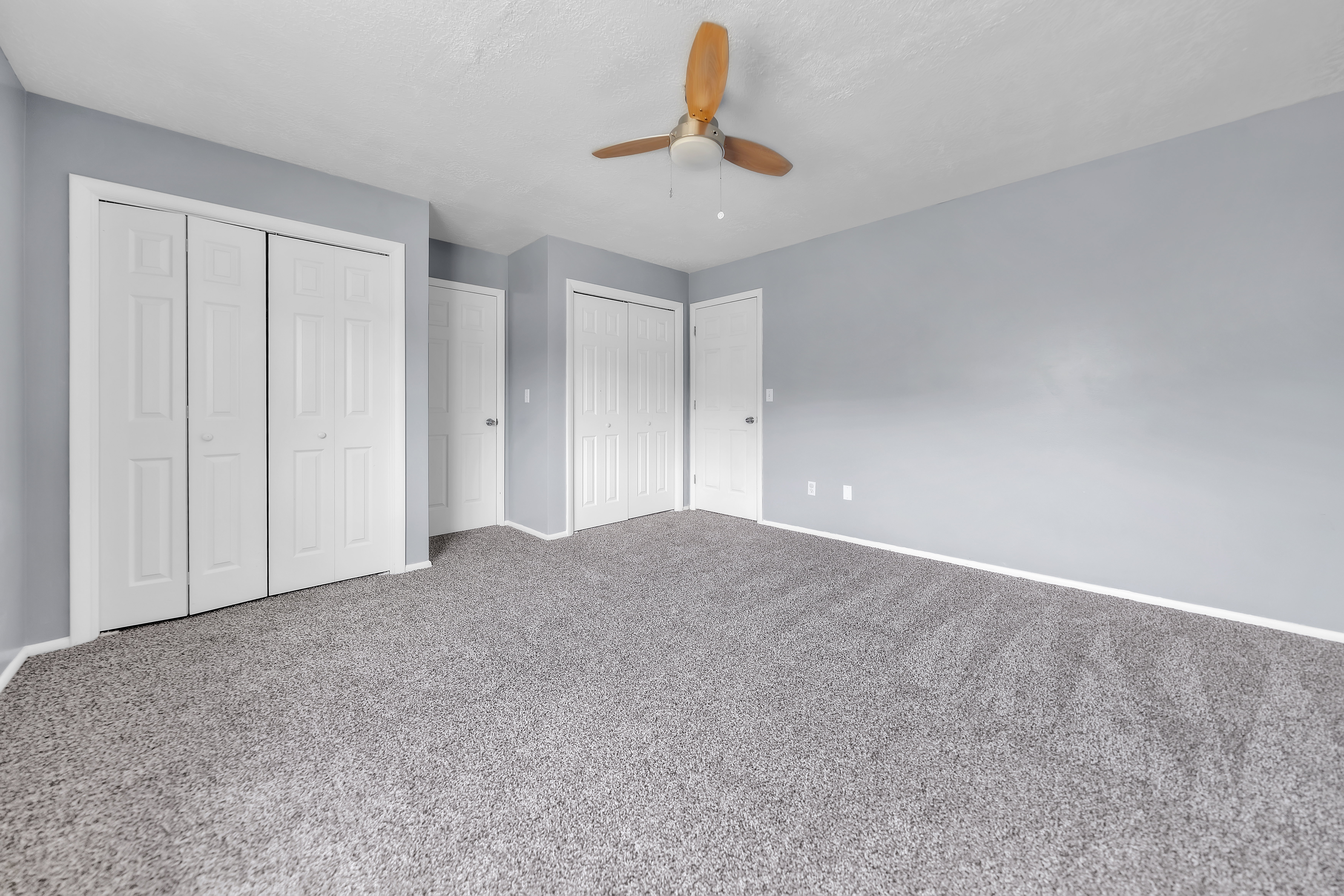Spacious room featuring light gray wall paint, gray carpet flooring, and a ceiling fan with wooden blades