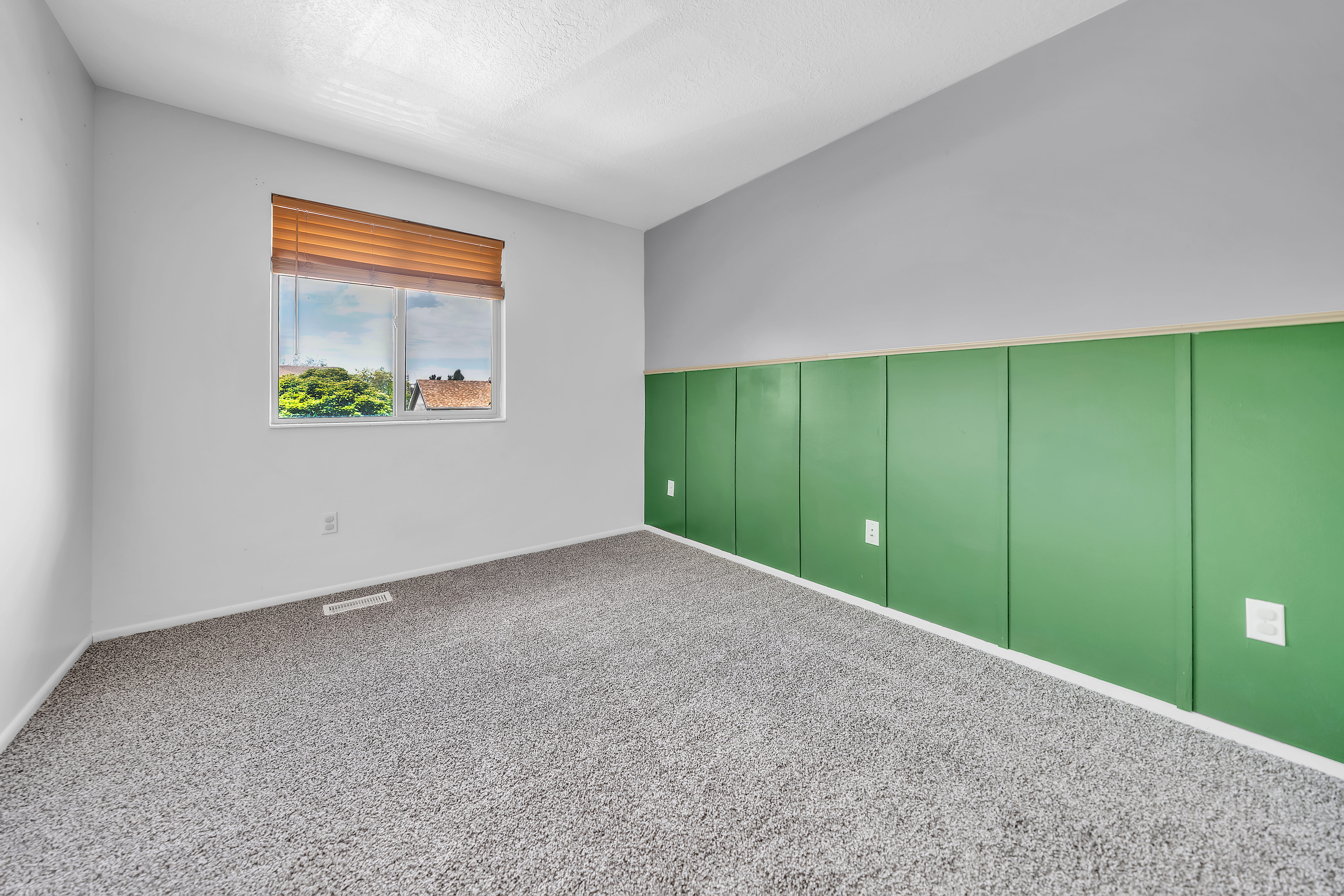 Room featuring a large window with a wood-finish blind, textured ceiling, light gray walls, and a green accent panel wall with white trim