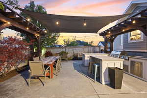 Patio terrace at dusk with an outdoor bar and dining area, exterior kitchen, a pergola, a patio, and a fenced backyard
