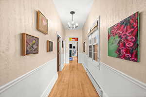 Corridor featuring a chandelier, light wood flooring, a baseboard heating unit, a wainscoted wall, and a textured wall