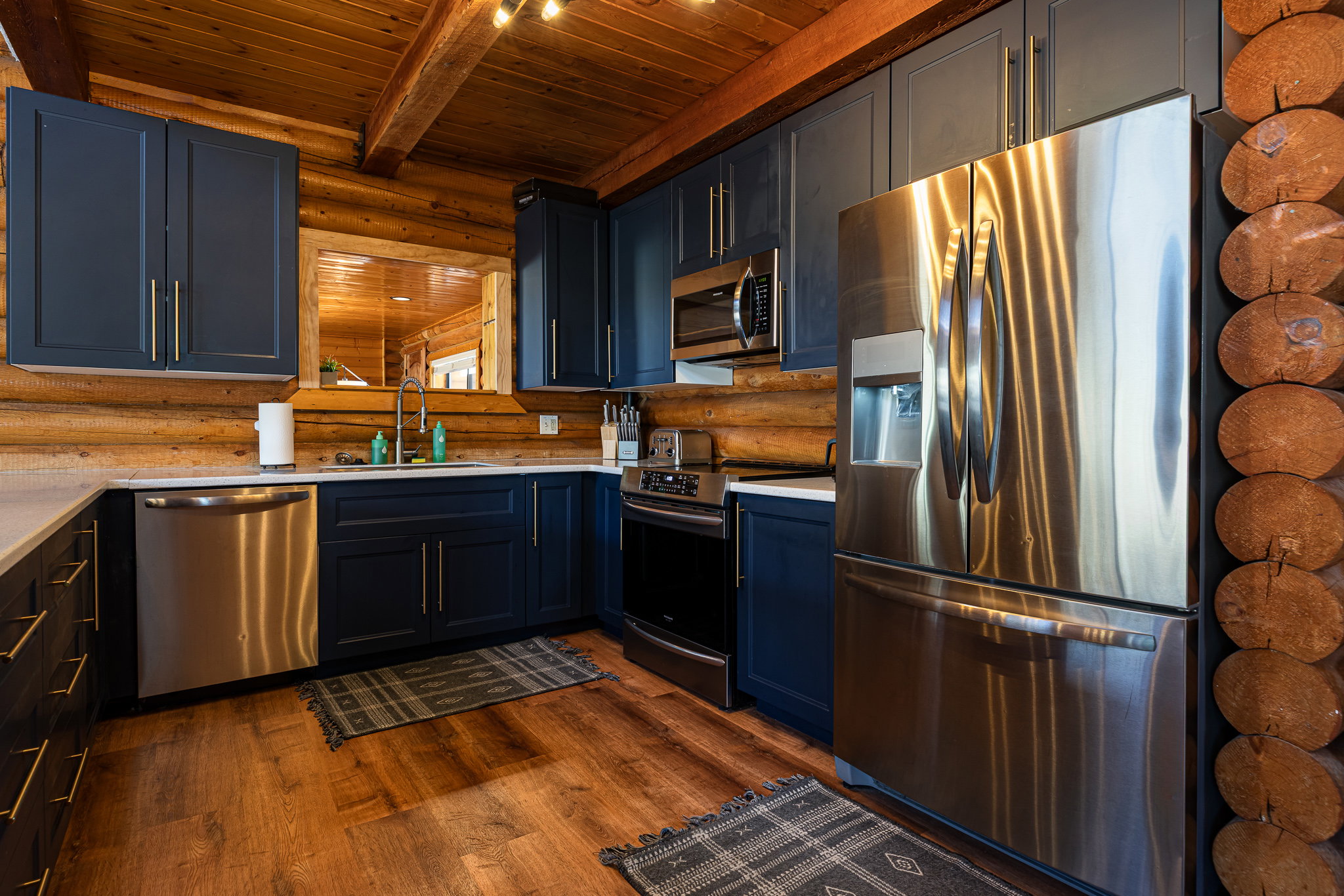 Kitchen with rustic walls, stainless steel appliances, dark wood-style flooring, a wooden ceiling with exposed beams, and light stone countertops