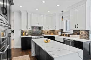 Two tone kitchen with dual tone cabinets, hanging light fixtures, light stone counters, dark wood-style floors, and stainless steel appliances