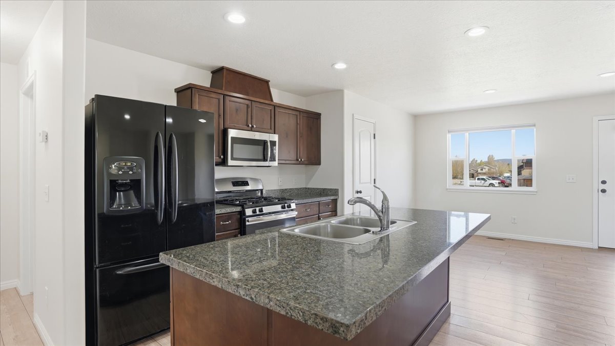 Kitchen featuring stainless steel appliances, dark wood finish cabinetry, light wood-type flooring, dark stone counters, and a center island with sink