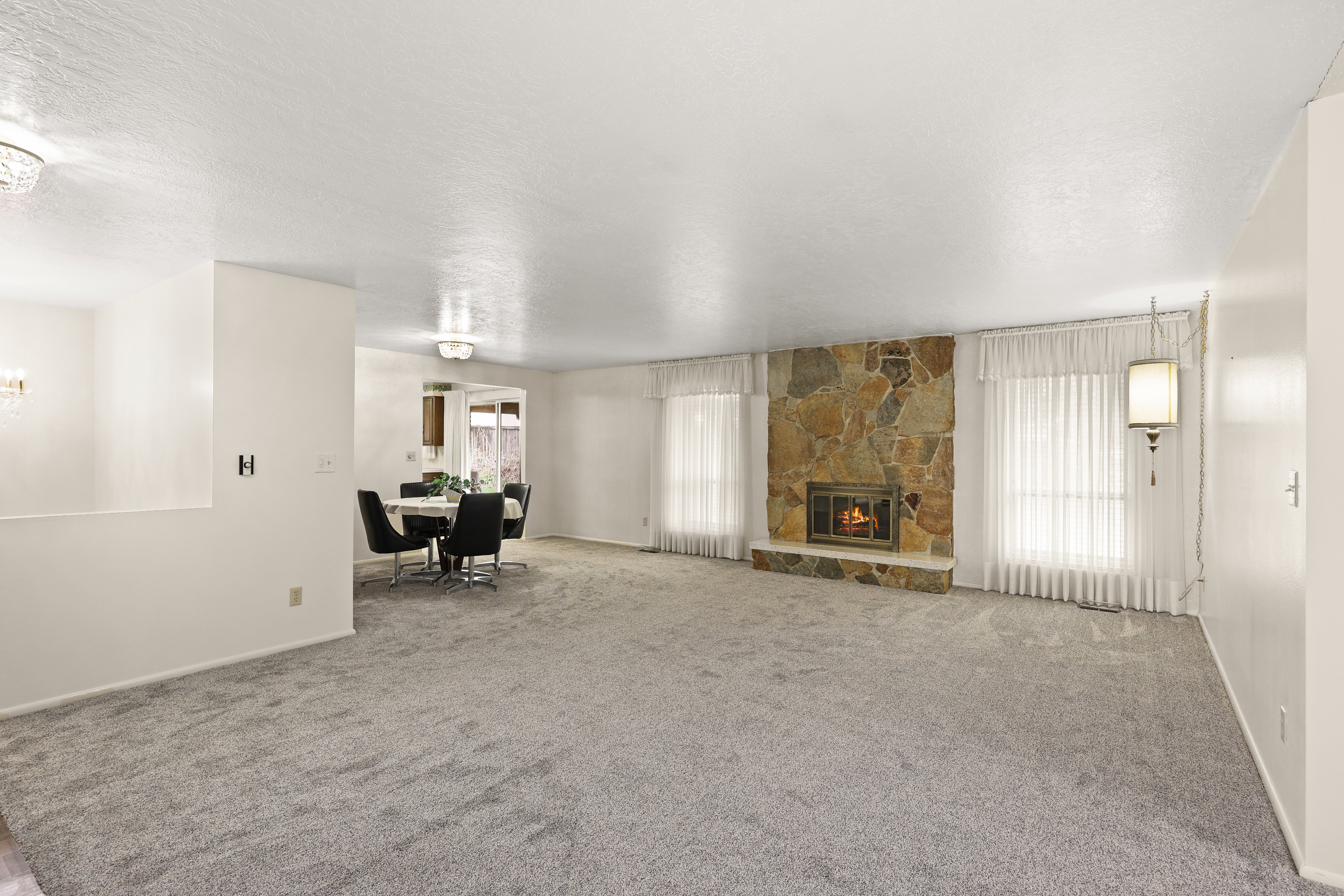 Spacious carpeted living area featuring a stone fireplace with a raised hearth, multiple windows with blinds, and a built-in pendant light fixture