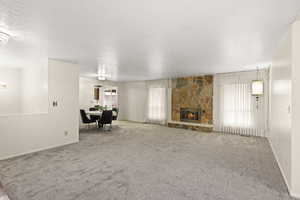 Spacious carpeted living area featuring a stone fireplace with a raised hearth, multiple windows with vertical blinds, and a built-in pendant light fixture