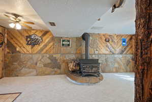 Unfurnished living room featuring a textured ceiling, a wood stove, carpet floors, ceiling fan, and wood walls