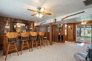 Dining area with ceiling fan, light colored carpet, a textured ceiling, wooden walls, and a dry bar