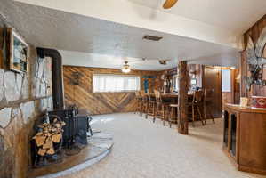 Dining space with a ceiling fan, wooden walls, light carpet, a textured ceiling, and a wood stove
