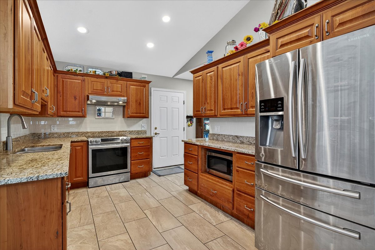 Kitchen with stainless steel appliances, wood finish cabinets, light stone countertops, vaulted ceiling, and tasteful backsplash