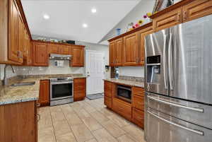 Kitchen with stainless steel appliances, wood finish cabinets, light stone countertops, vaulted ceiling, and tasteful backsplash