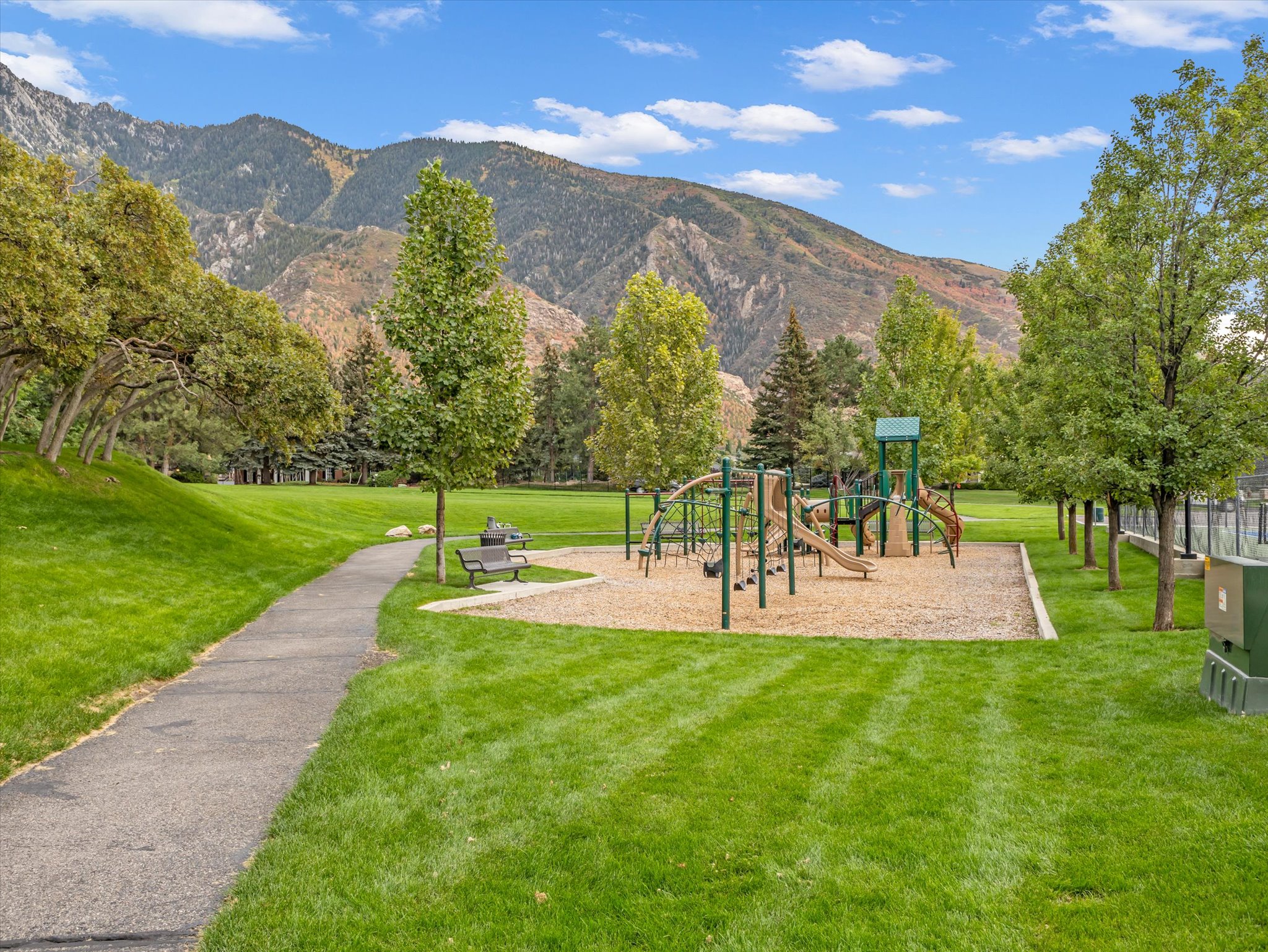 Expansive park area featuring a playground with slides and climbing structures, a paved walking path, mature trees, and a mountain backdrop