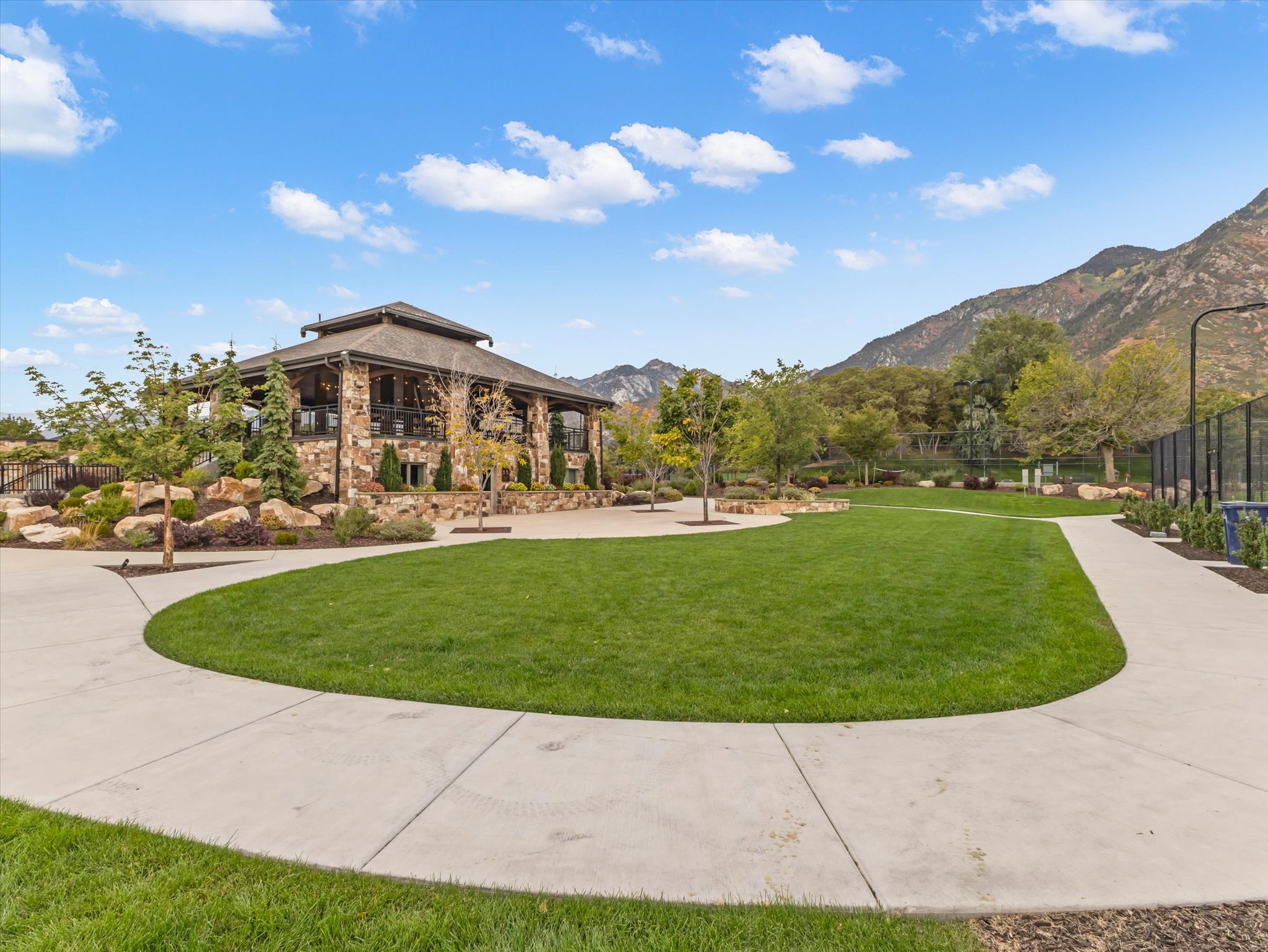 Expansive green lawn surrounded by a concrete pathway and mature trees, featuring a stone-clad building with a covered patio and a mountain backdrop