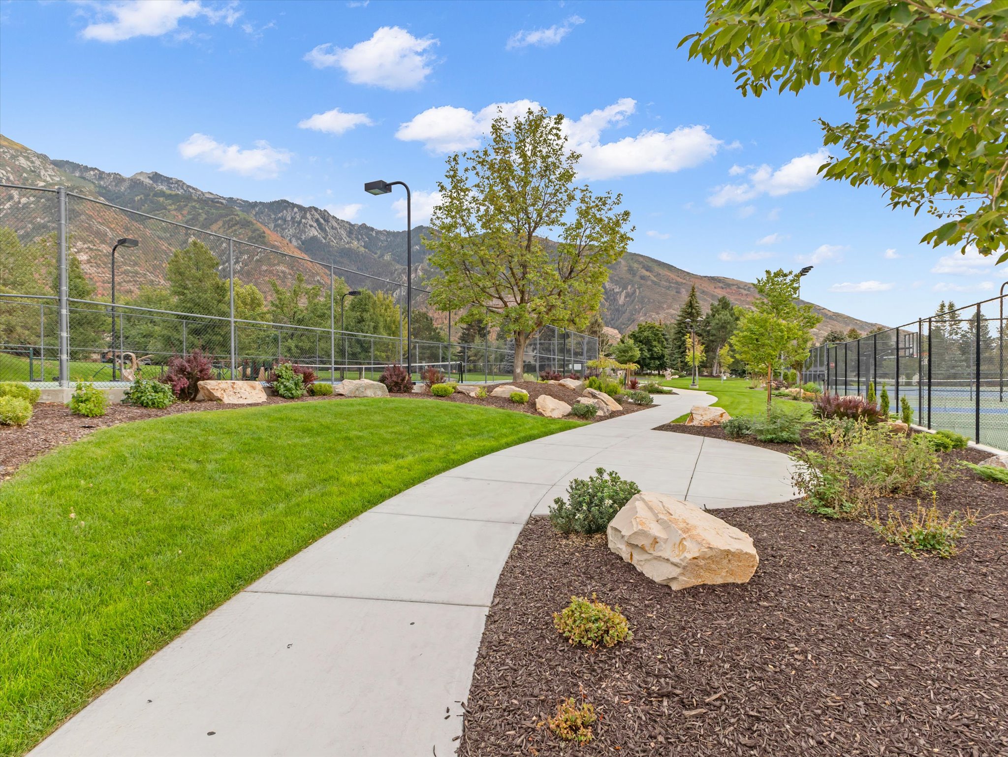 Winding paved pathway through landscaped grounds, featuring mature trees, ornamental shrubs, and decorative boulders