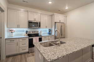 Kitchen featuring stainless steel appliances, light stone countertops, decorative backsplash, light wood-style floors, and a center island with sink